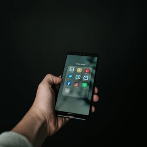 Close-up of a hand holding a smartphone displaying various social media app icons on a dark background.