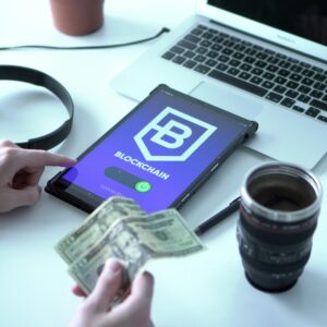 Tablet with blockchain app, laptop, and dollar bills on desk depicting digital finance.